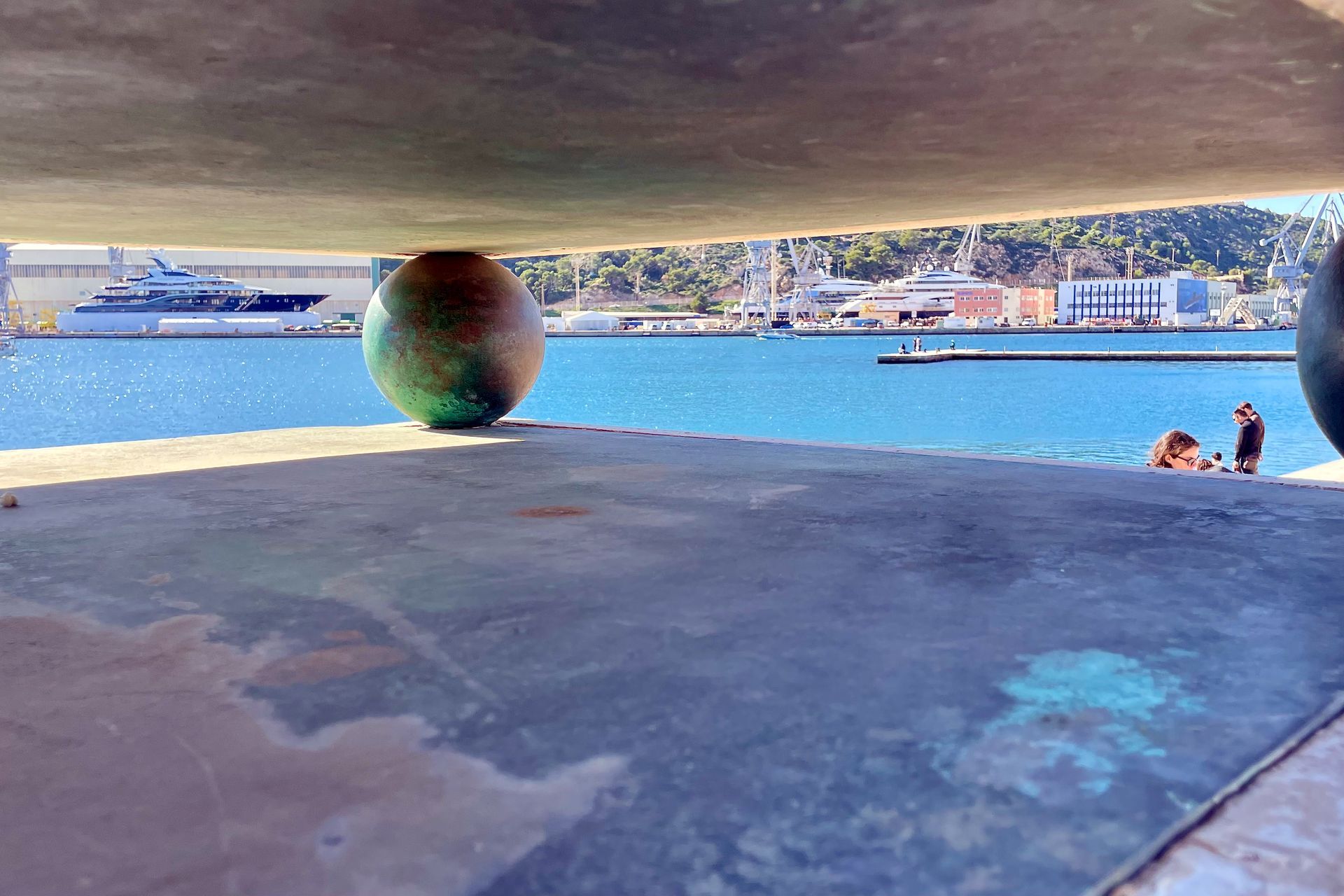 Gezicht onder een bouwwerk, met een haven met een boot, gebouwen en twee mensen.