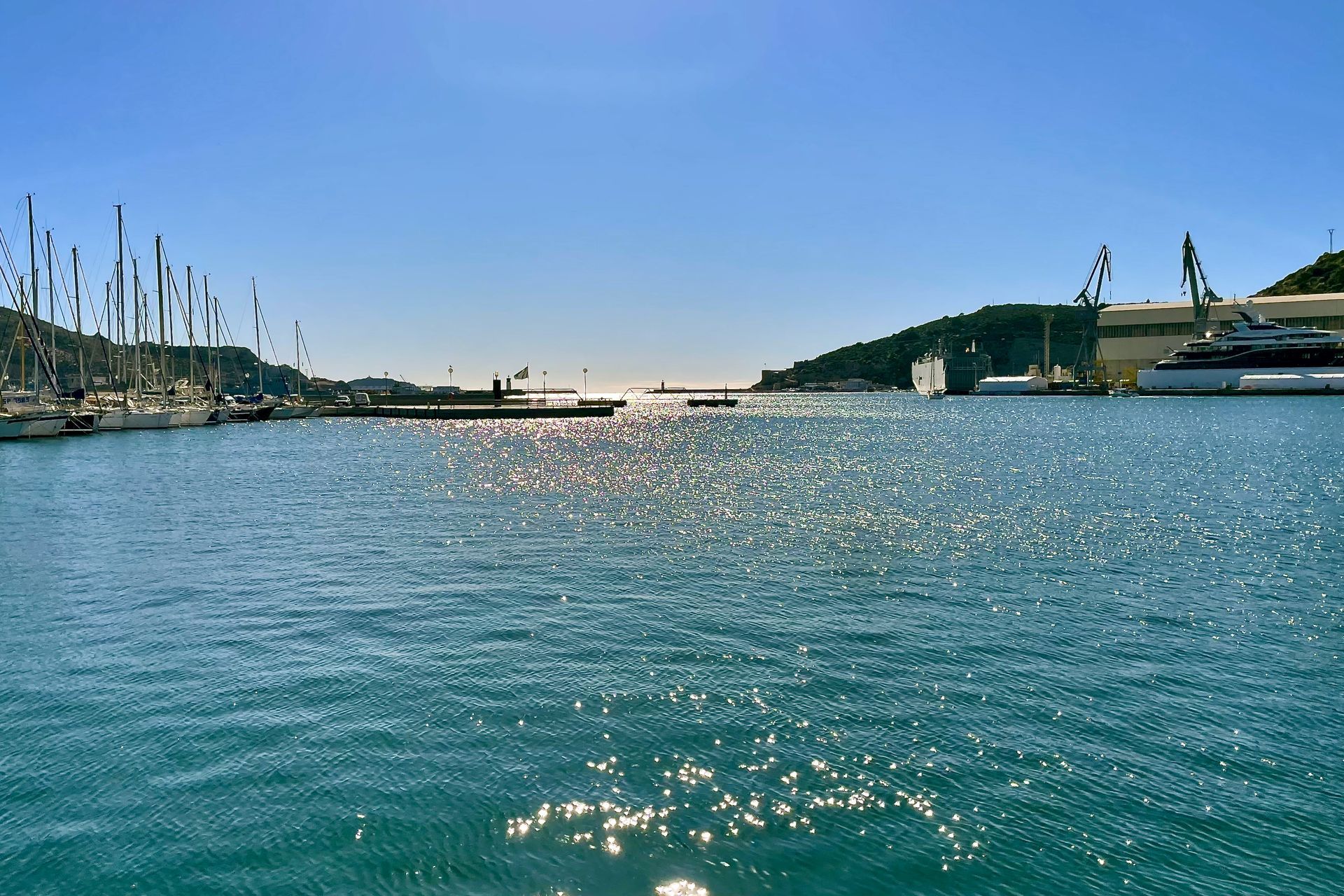 Fel zonlicht schittert op het water van een haven, met zeilboten aan de linkerkant en gebouwen/kranen aan de rechterkant, onder een strakblauwe lucht.