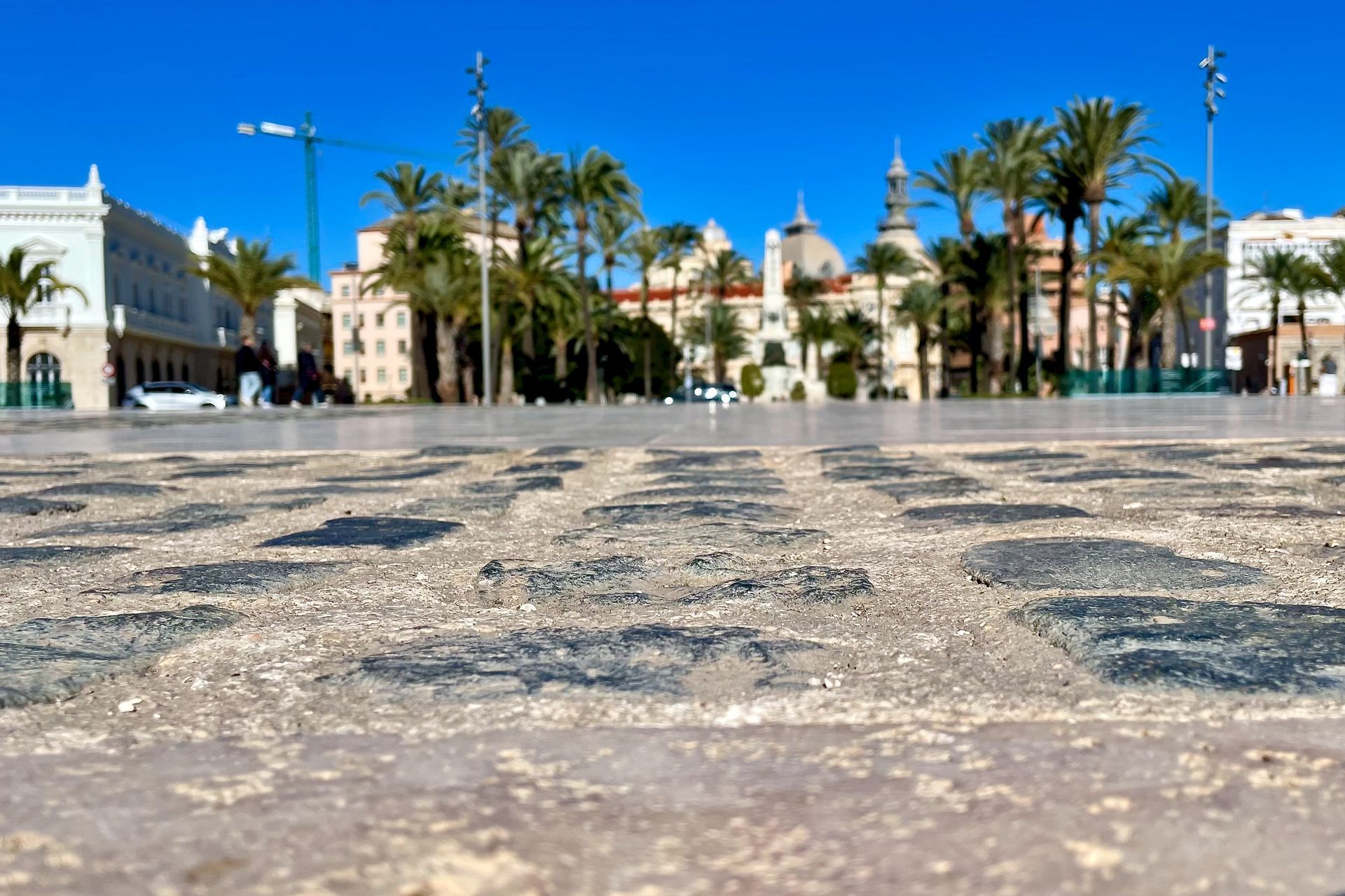 Voorgrond met kasseien; gebouwen en palmbomen tegen een blauwe lucht op een stadsplein.