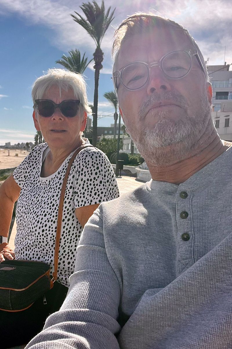 Man en vrouw poseren buiten; man neemt selfie. Palmbomen en strand op de achtergrond. Fel zonlicht.