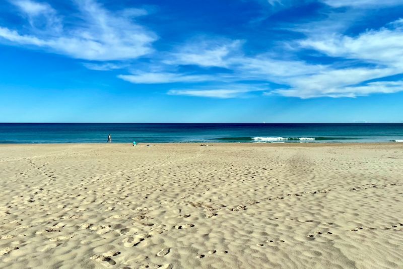 Zandstrand onder een helderblauwe lucht met witte wolken, met uitzicht op de oceaan.