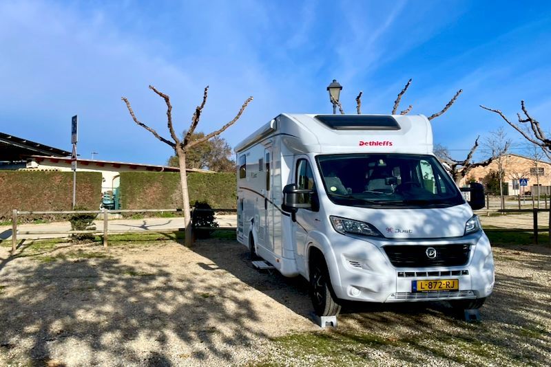 Witte camper geparkeerd op grind op een zonnige camping met kale bomen en een blauwe lucht.