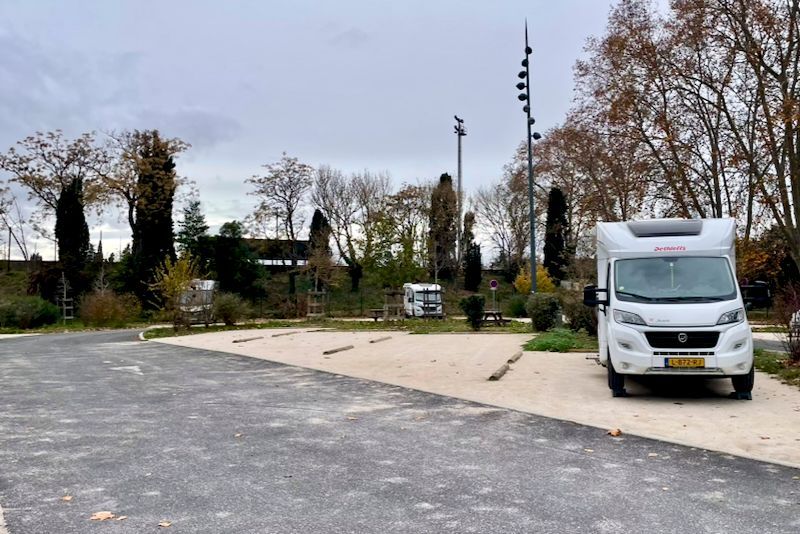 Camper geparkeerd op een kampeerterrein met grindpaden. Bomen en een bewolkte lucht op de achtergrond.