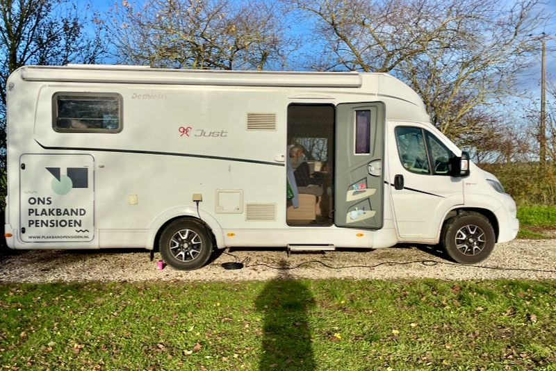 Witte camper geparkeerd op een onverharde weg, deur open. Er is iemand binnenin te zien. Groen gras en bomen op de achtergrond.