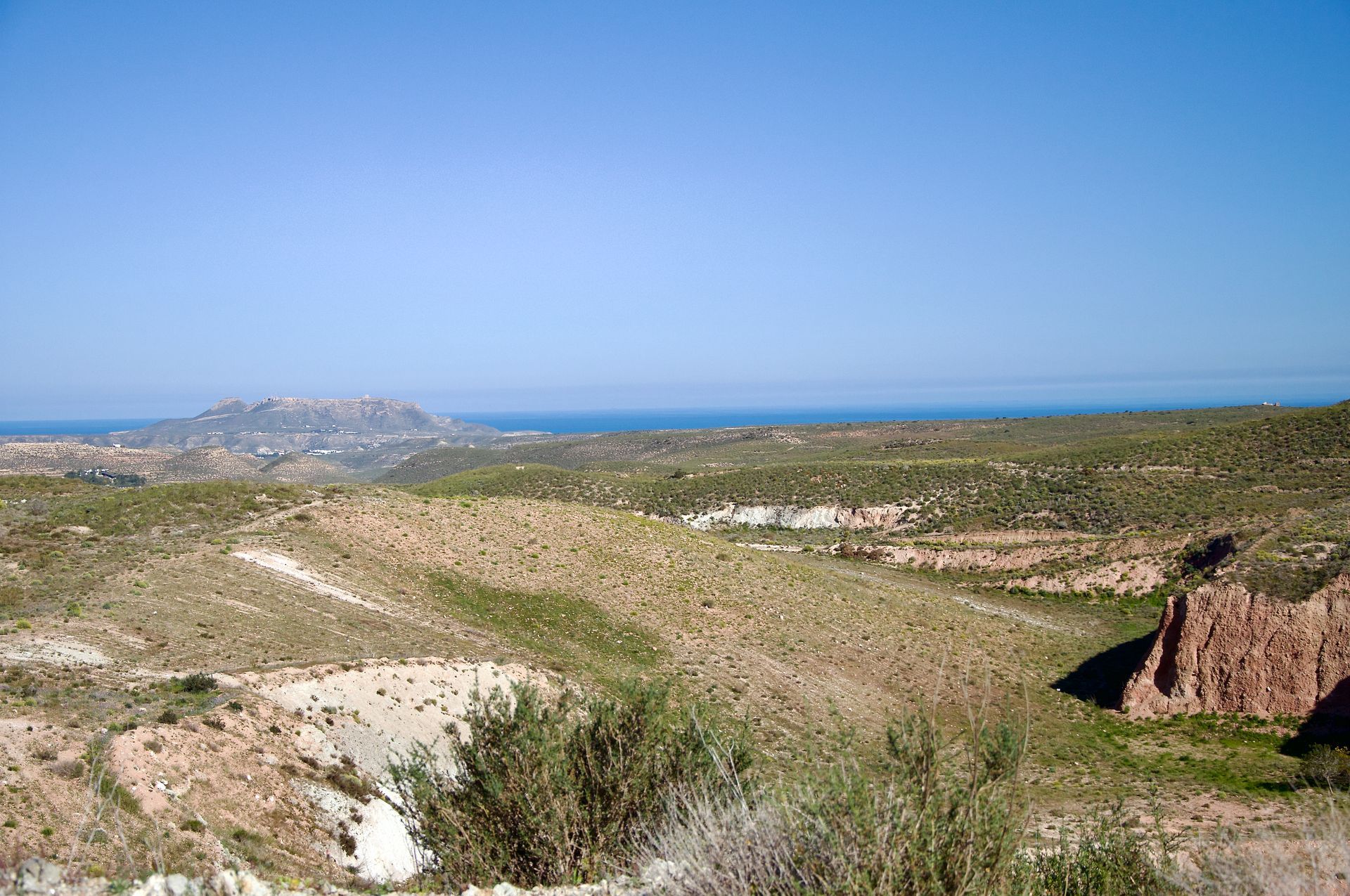 Een uitgestrekt, dor, heuvelachtig landschap met struikgewas onder een helderblauwe hemel, met uitzicht op een verre zee.