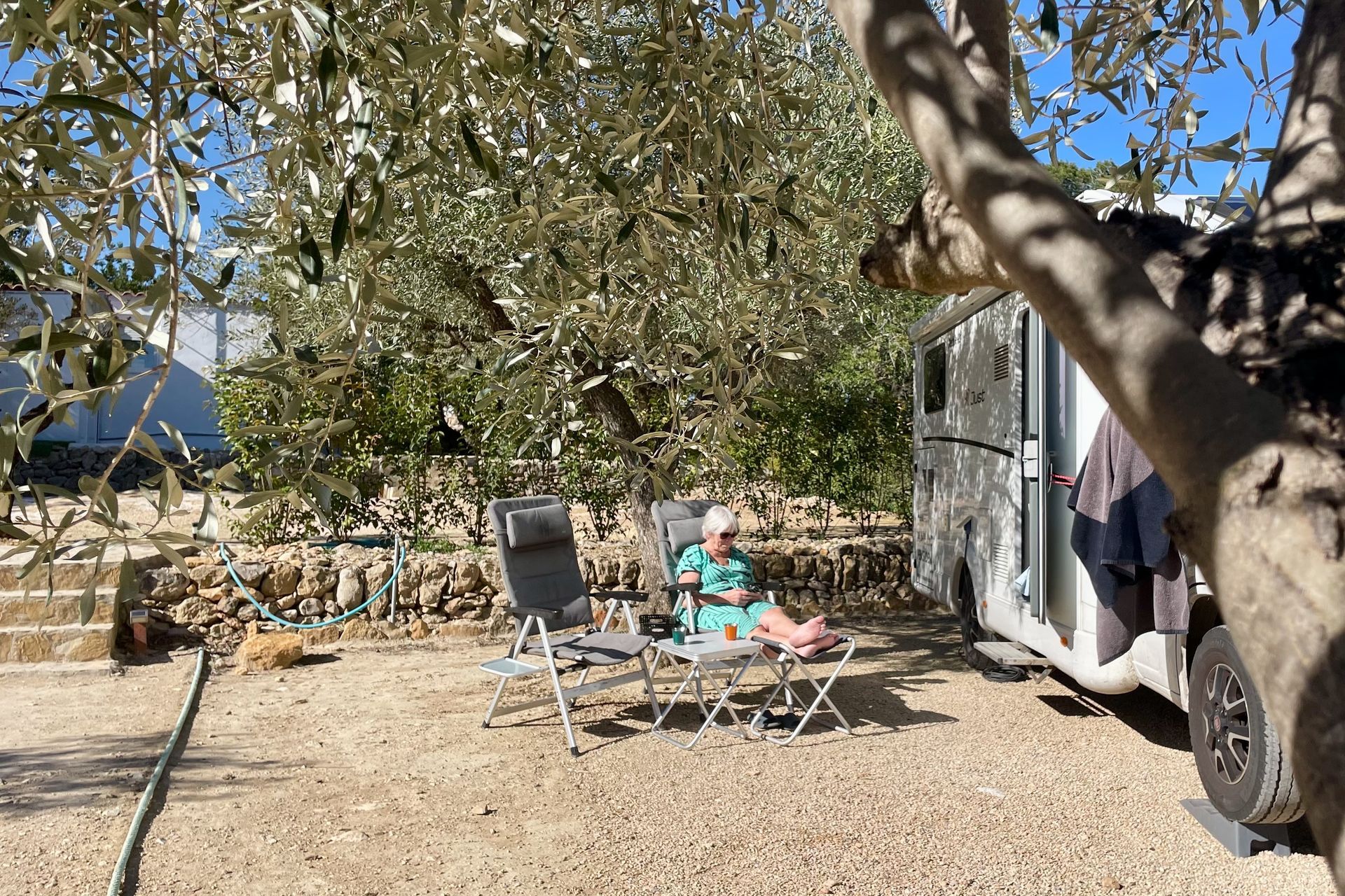 Een persoon zit op een stoel bij een camper onder olijfbomen. Op de achtergrond is de grond droog en de lucht blauw.