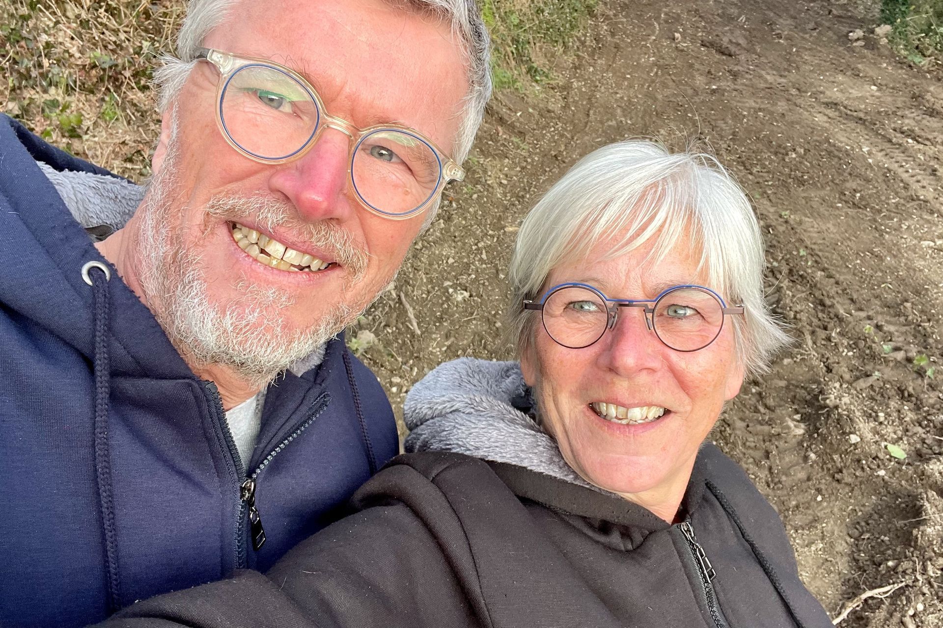 Jut en Jul Een stel in hoodies lacht buiten, de een met een grijze baard en bril, de ander met grijs haar en bril.