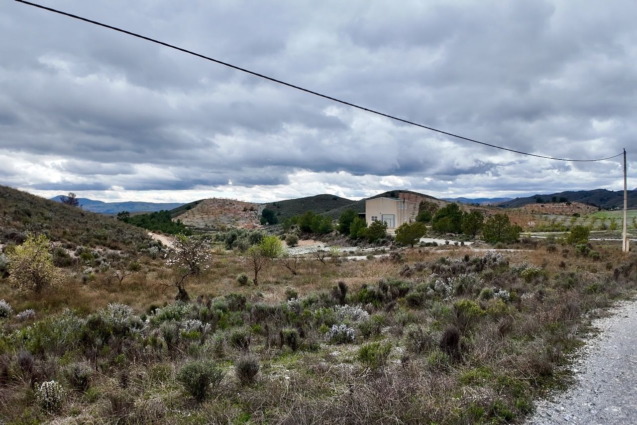 Een grindpad leidt naar een klein gebouw, verscholen tussen droge, met struiken begroeide heuvels onder een bewolkte hemel.