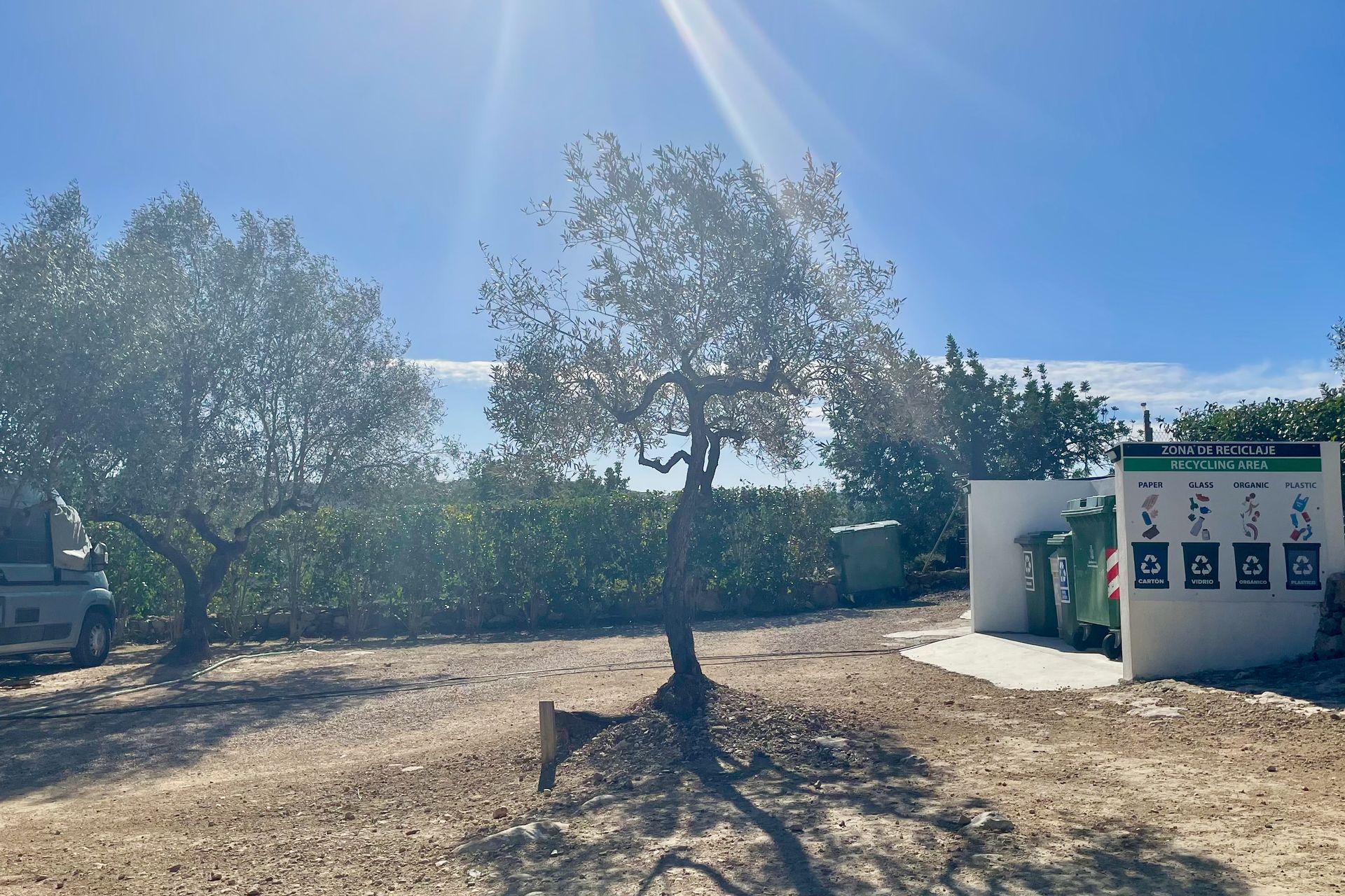 Bomen, een recyclingcentrum met groene containers en borden, onder een helderblauwe hemel.
