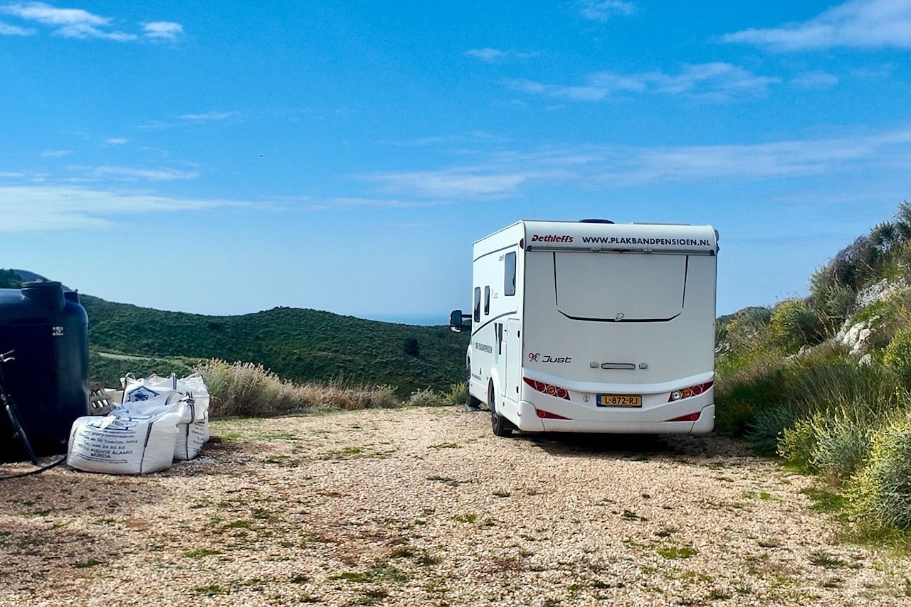 Een witte camper staat geparkeerd op een grindheuvel met uitzicht op glooiende groene heuvels en de oceaan in de verte, onder een blauwe hemel.