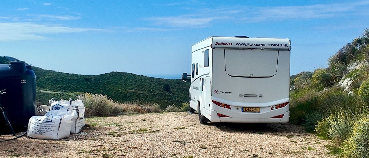 Witte camper geparkeerd op een grindpad naast struiken en heuvels onder een blauwe hemel.