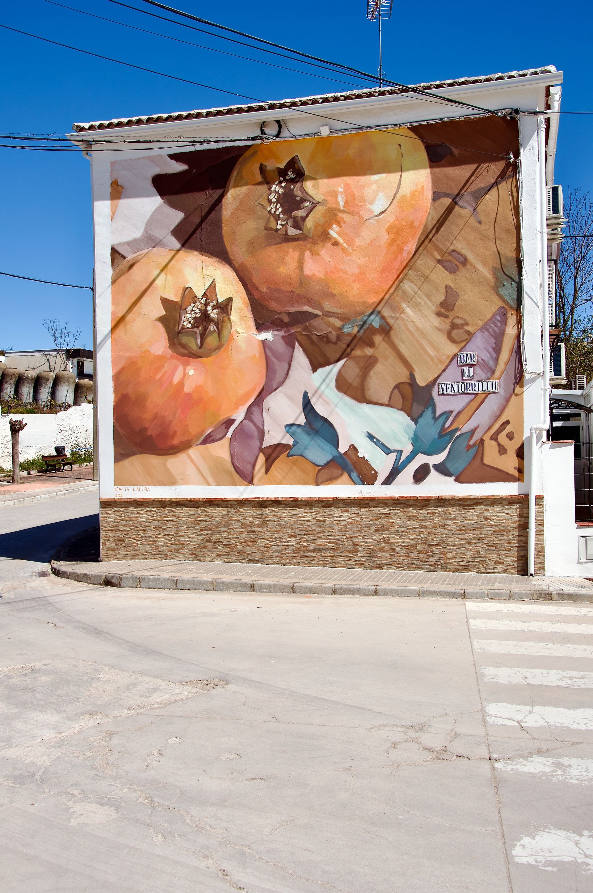 Een muurschildering van twee grote, lichtoranje granaatappels op de buitenmuur van een wit gebouw onder een helderblauwe hemel.