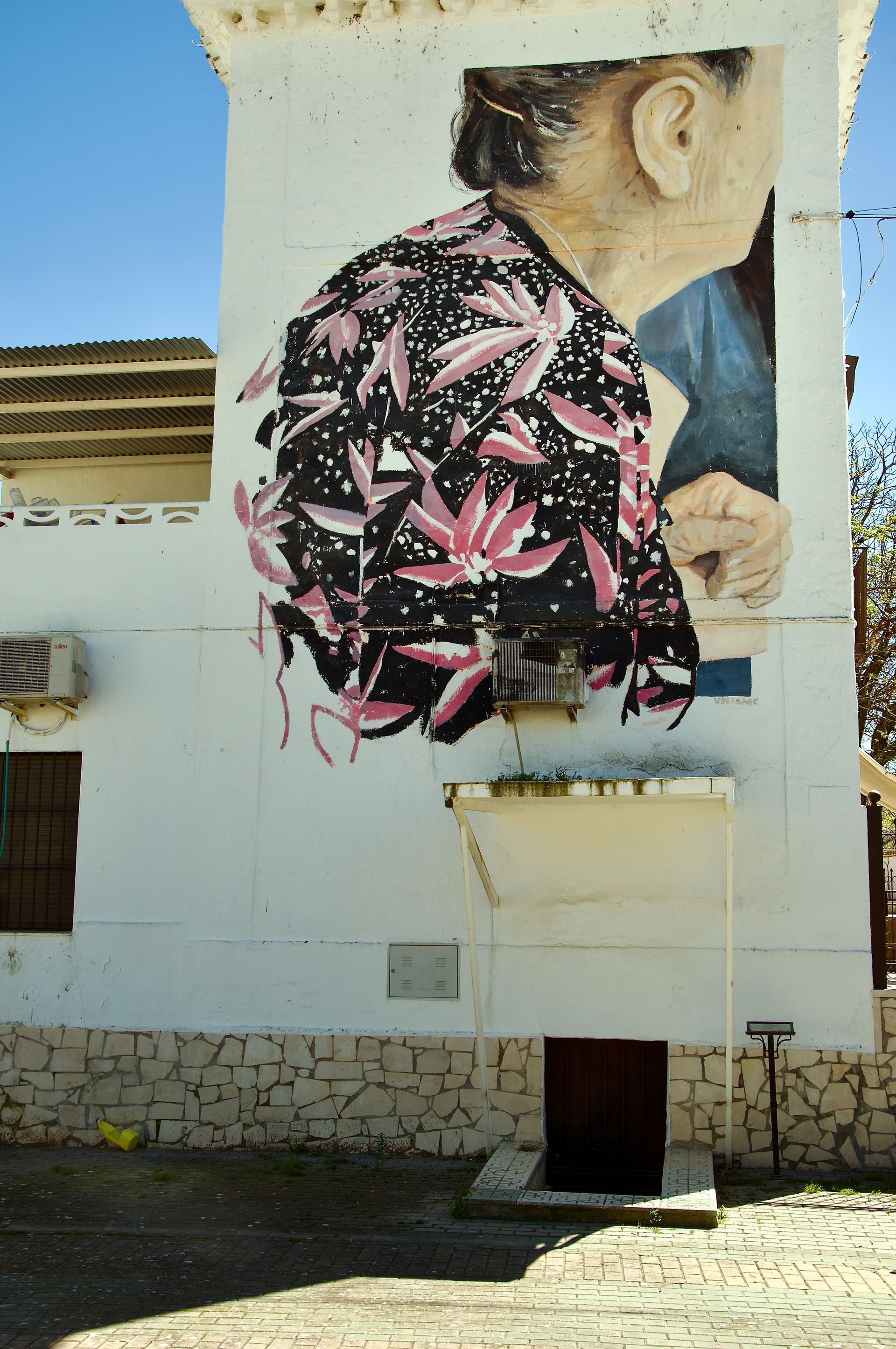 Op een muurschilding op een wit gebouw is een persoon te zien die een zwart shirt met een roze bloemenprint draagt.