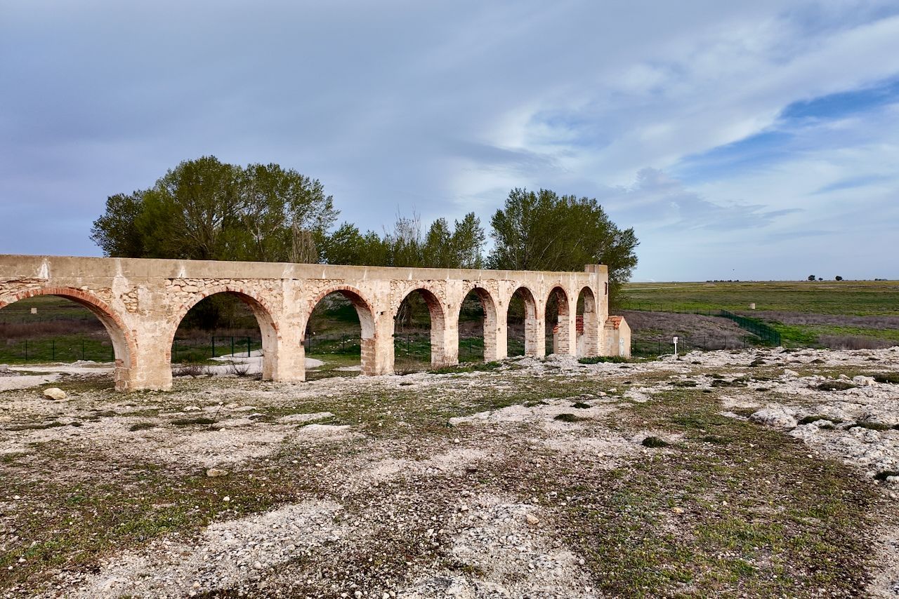 Een verweerd, meerboogvormig stenen aquaduct strekt zich uit over een droog, rotsachtig veld onder een gedeeltelijk bewolkte hemel.