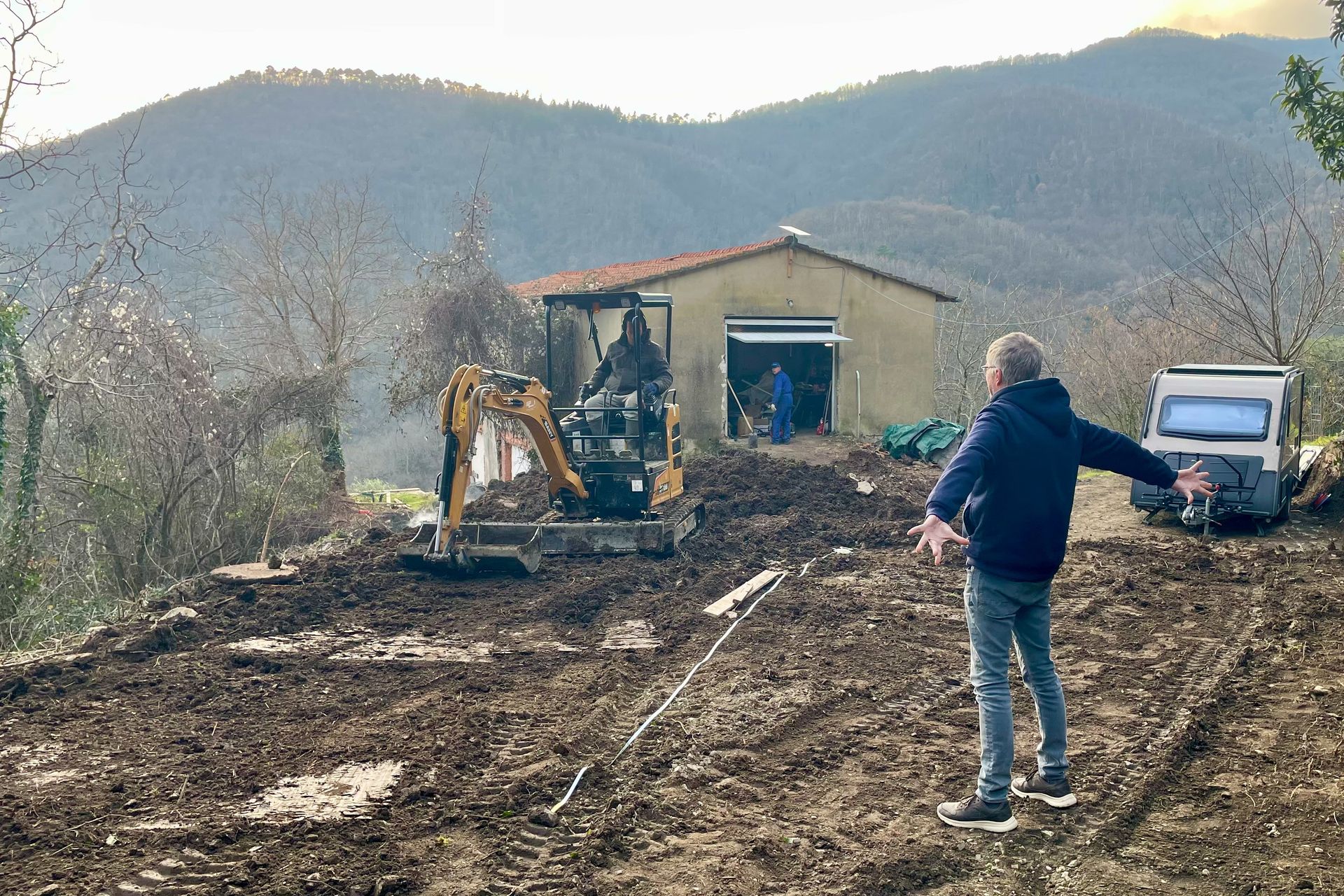 Nog even egaliseren Een man wijst naar een bouwterrein met een graafmachine, een loods en bergen op de achtergrond.