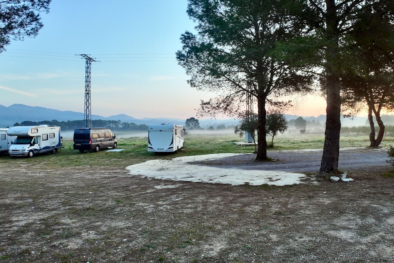 Een camping met campers en bomen bij zonsopgang, met een mistig veld en bergen in de verte.