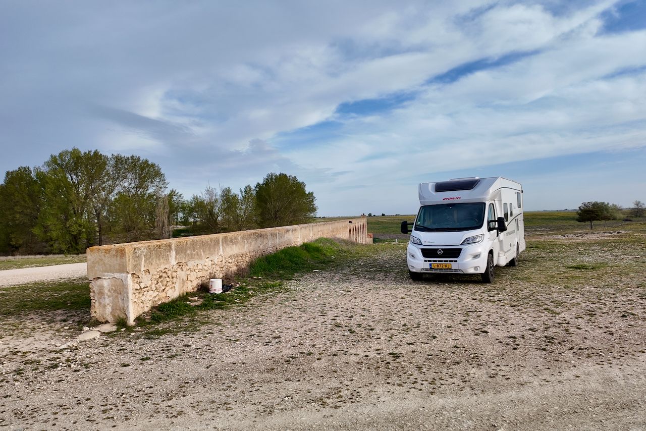 Een witte camper staat geparkeerd op een grindterrein naast een lage, verweerde stenen muur met bomen onder een blauwe, bewolkte hemel.
