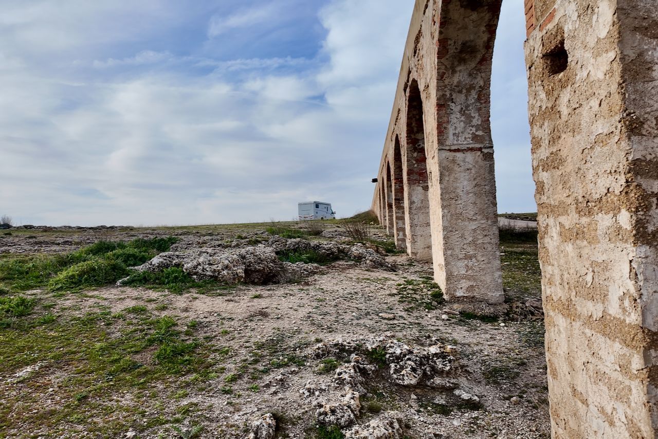 Een reeks verweerde stenen bogen van een vervallen aquaduct strekt zich uit over een rotsachtig, grasrijk landschap onder een bewolkte hemel.