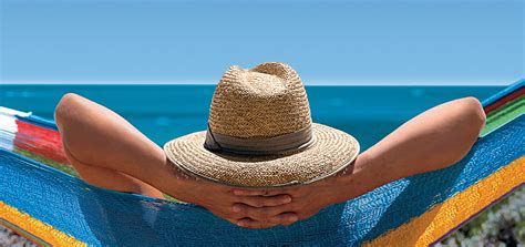 Person relaxing in a colorful hammock by the ocean, wearing a straw hat with arms behind head