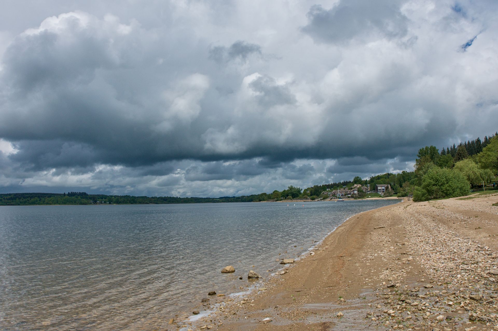 Een bewolkte oever met kalm water, een zandstrand en bomen langs de verre oever.