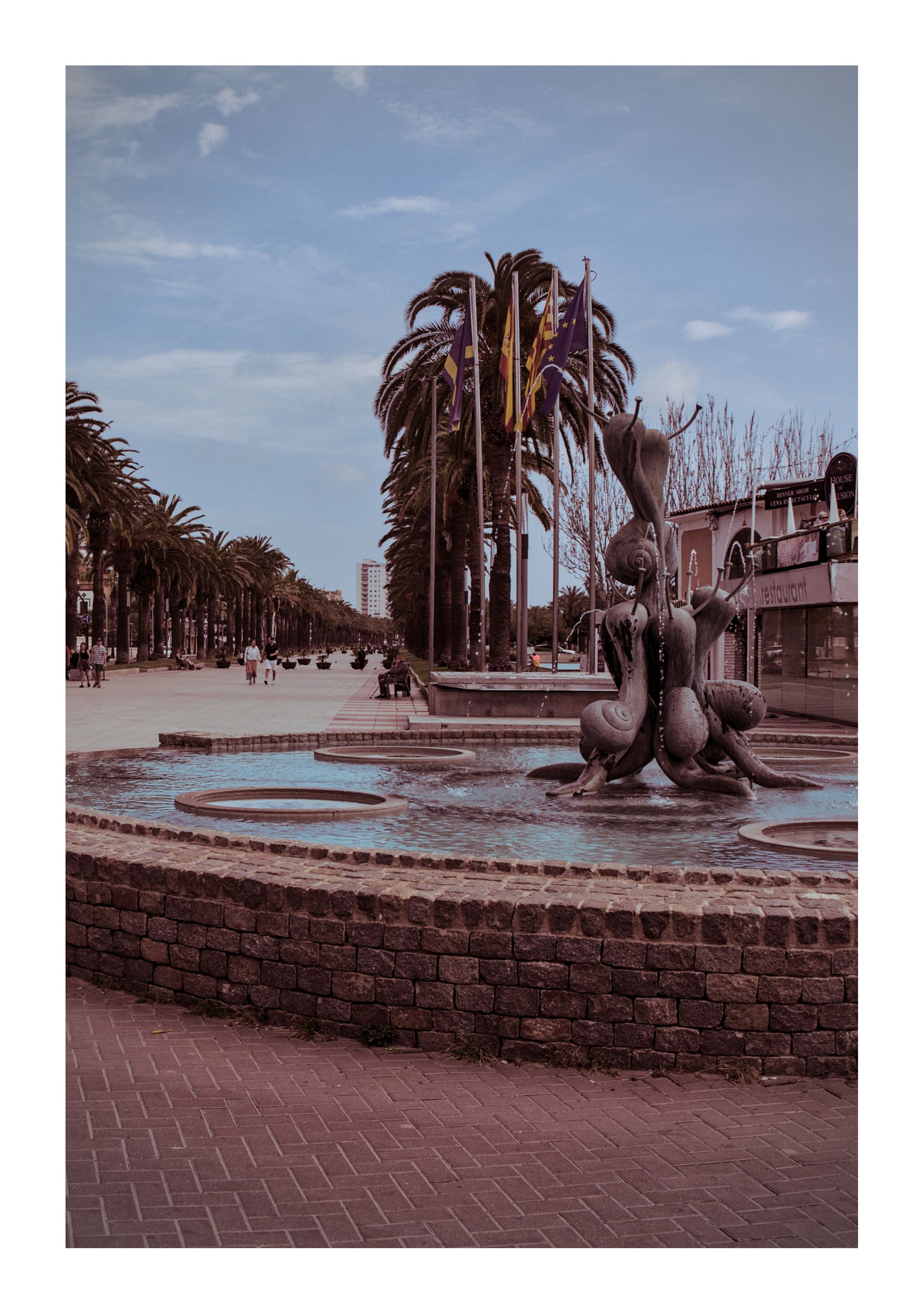 Een met palmbomen omzoomd plein met een ronde fontein en een stenen sculptuur onder een bewolkte hemel.