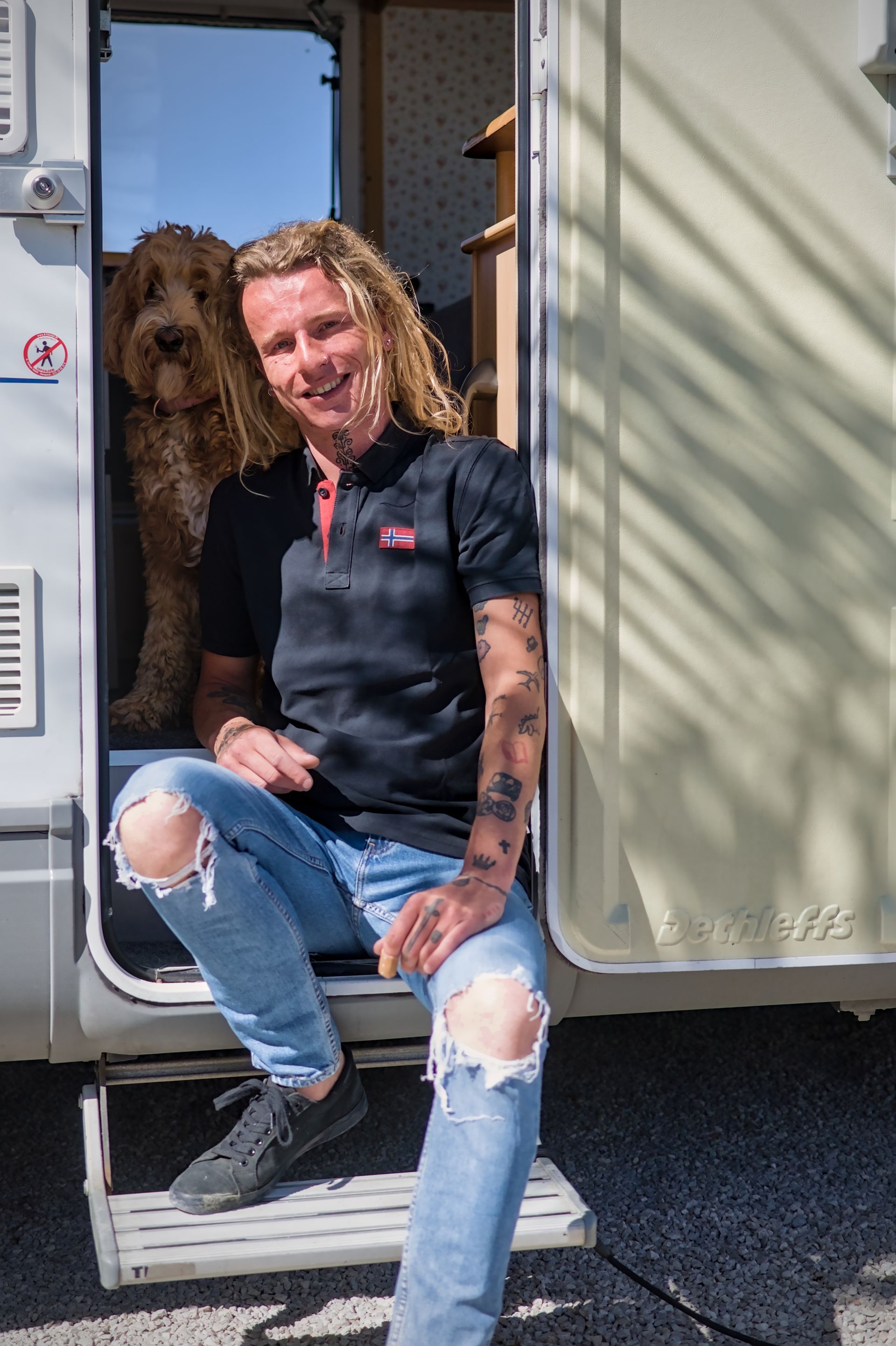 Een persoon zit in de deuropening van een camper, gekleed in een zwart shirt en gescheurde jeans, met een hond achter zich.