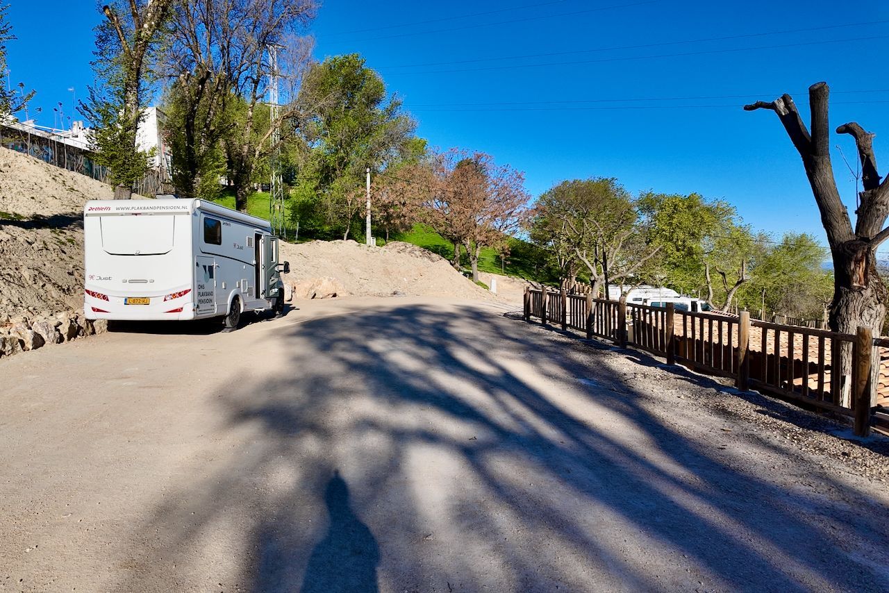 Een witte camper staat geparkeerd op een geasfalteerde parkeerplaats naast een houten hek, met bomen en een helderblauwe lucht op de achtergrond.