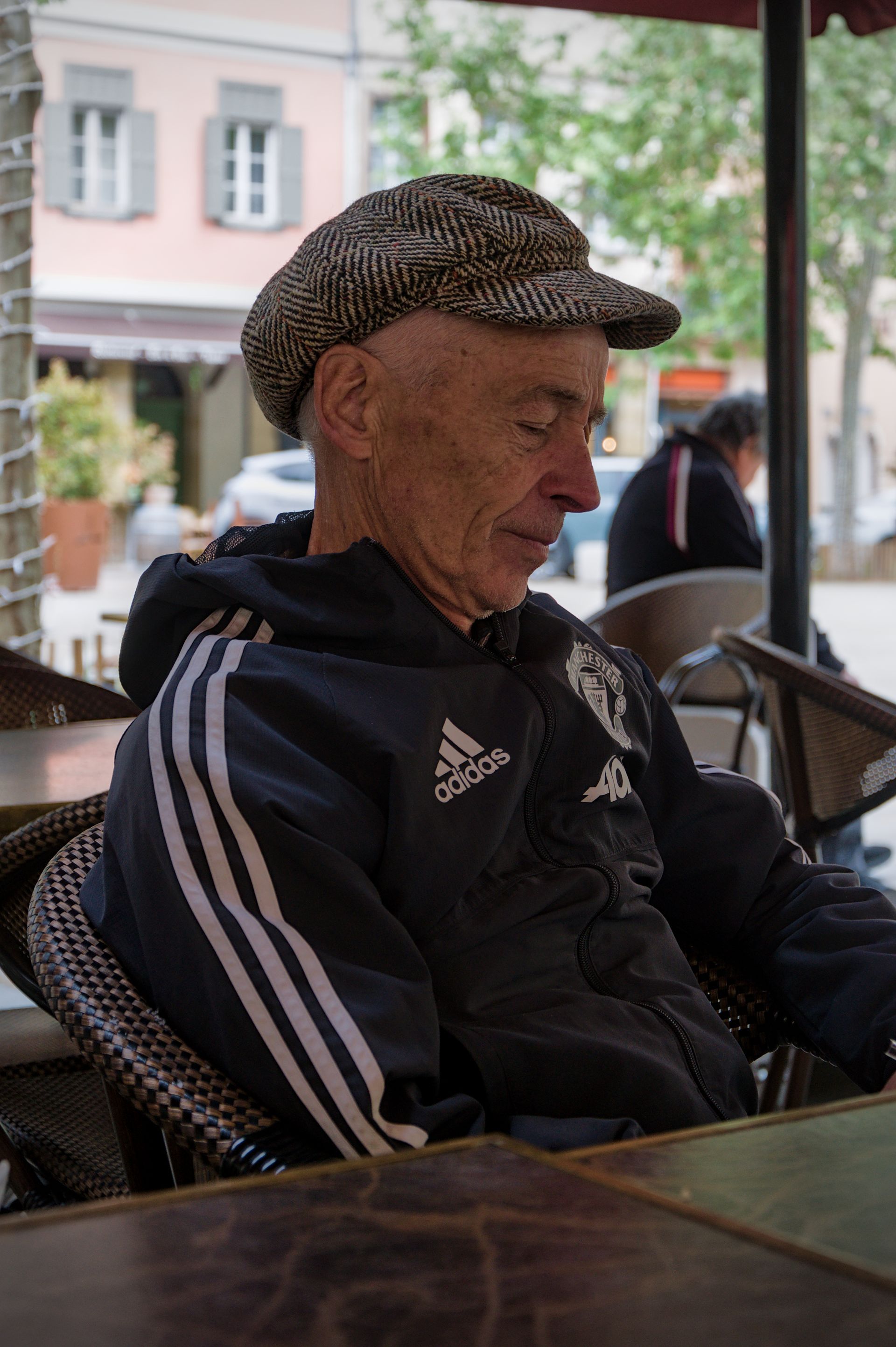 Een man in een zwart trainingsjack en een geruite pet zit aan een tafeltje op een terras.