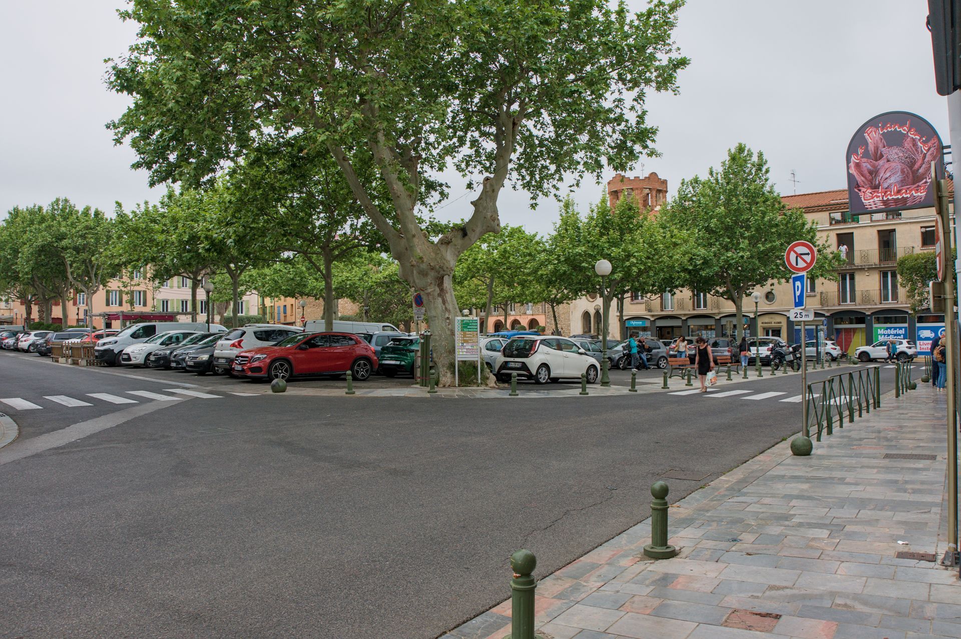 Een met bomen omzoomd stadsplein met geparkeerde auto's, voetgangers en een stenen trottoir in de buurt van winkels en een groot uithangbord.