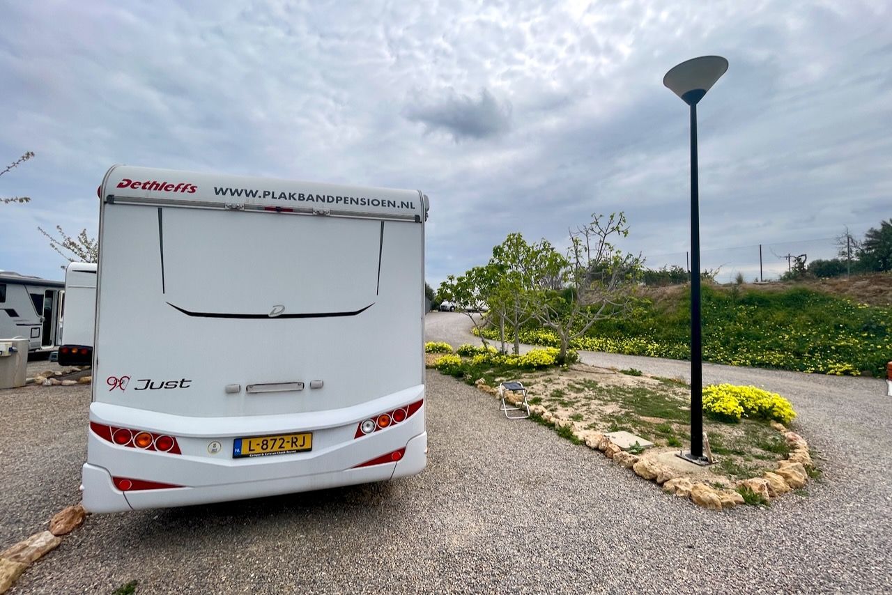 Achteraanzicht van een witte camper geparkeerd op een grindterrein bij een lantaarnpaal onder een bewolkte hemel.