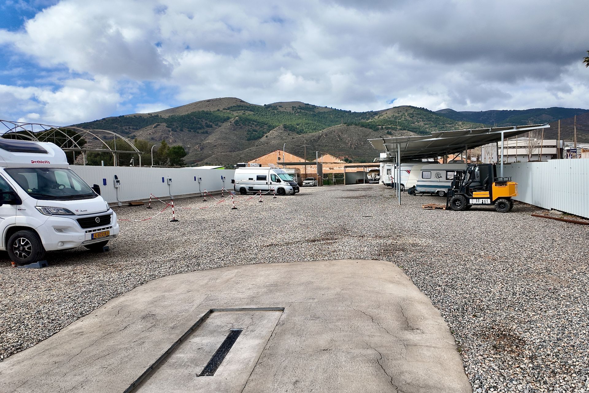 Een witte camper staat geparkeerd op een grindterrein vlakbij een betonnen afwateringsgebied, met bergen, gebouwen en een heftruck achterin.