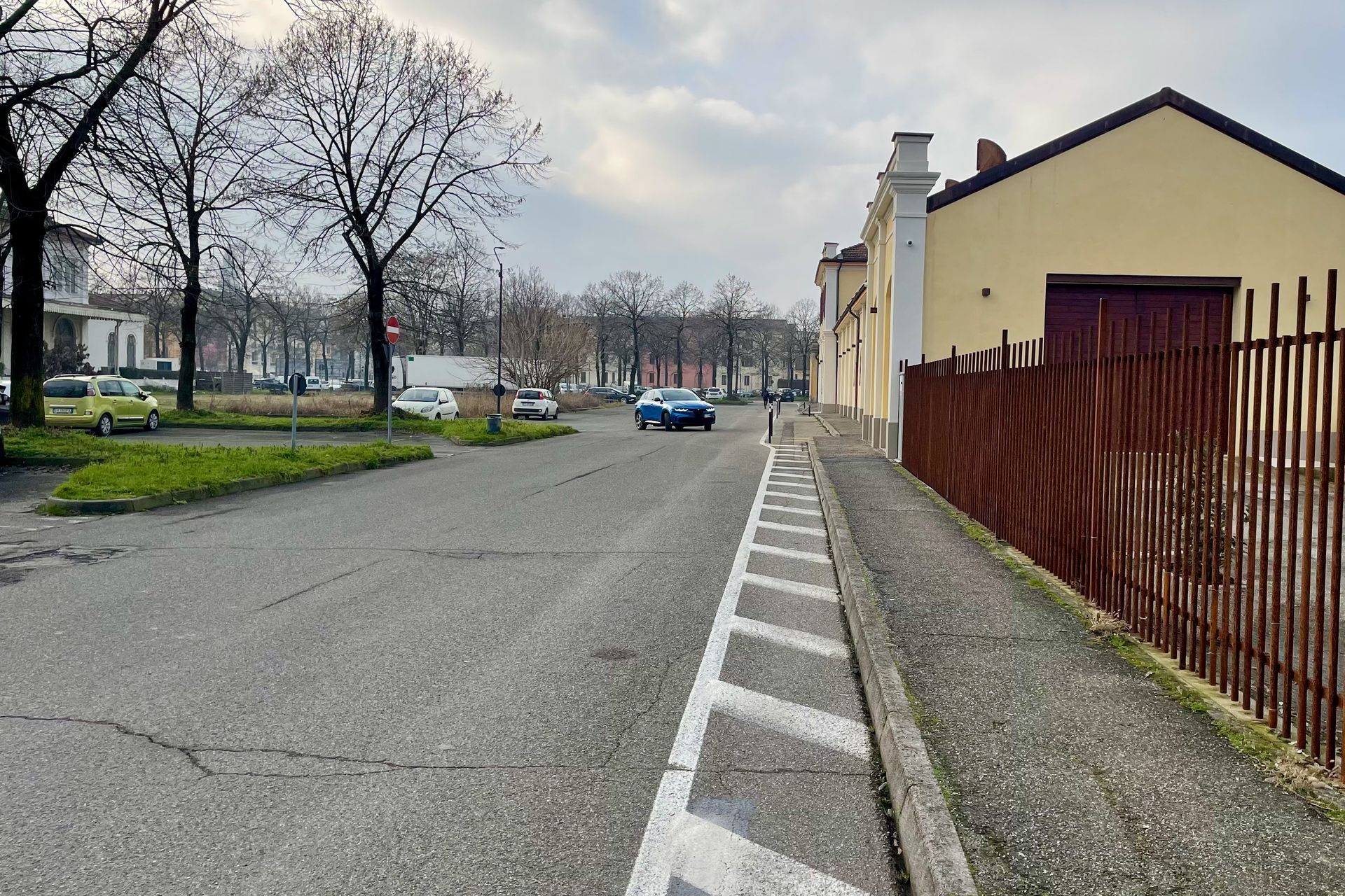 Straatbeeld met gebouwen, bomen en een blauwe auto op de weg. Lichtgekleurde stoeprand en hek op de voorgrond.