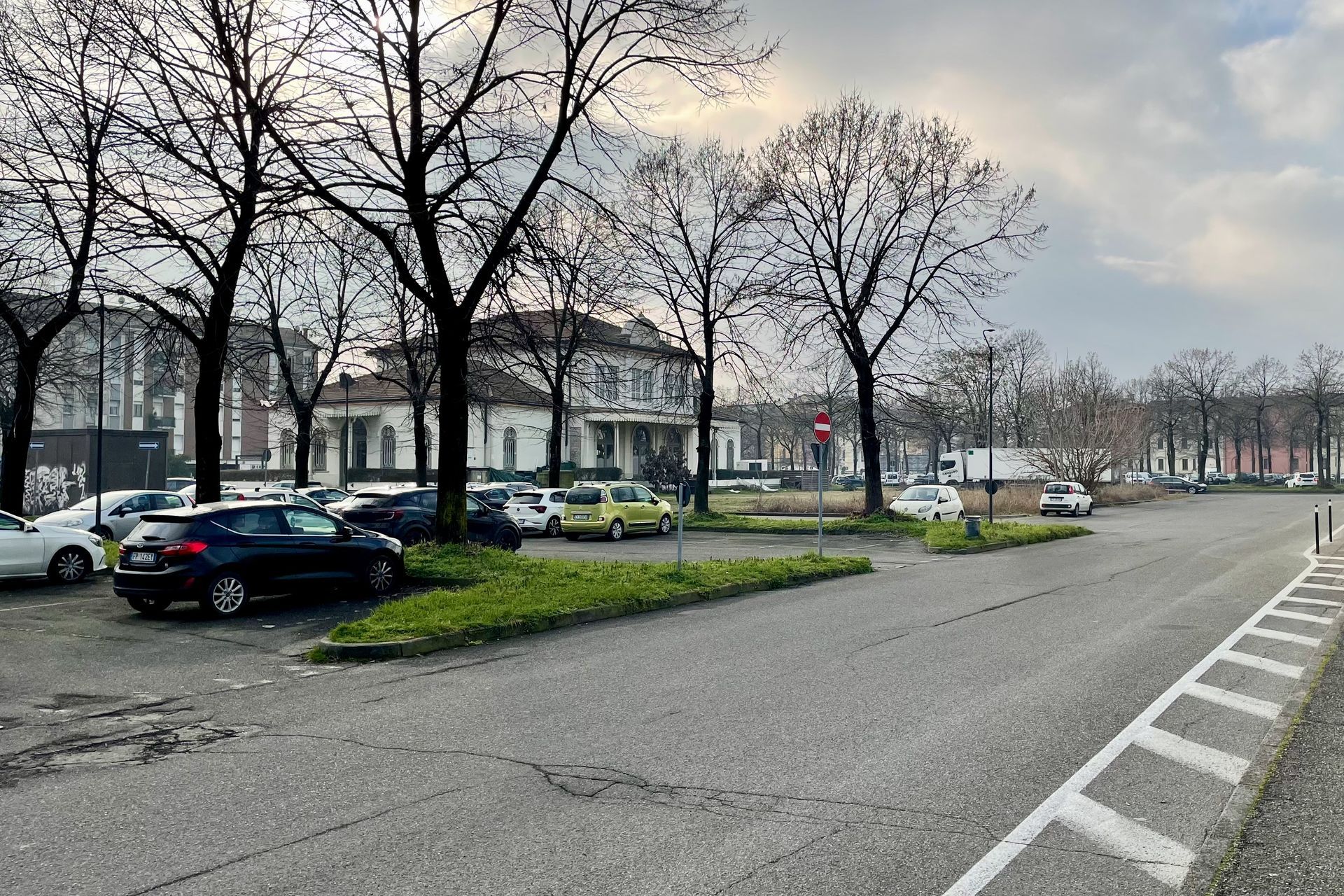 Auto's geparkeerd op een verhard terrein met bomen, een gebouw en een weg op de achtergrond, op een bewolkte dag.