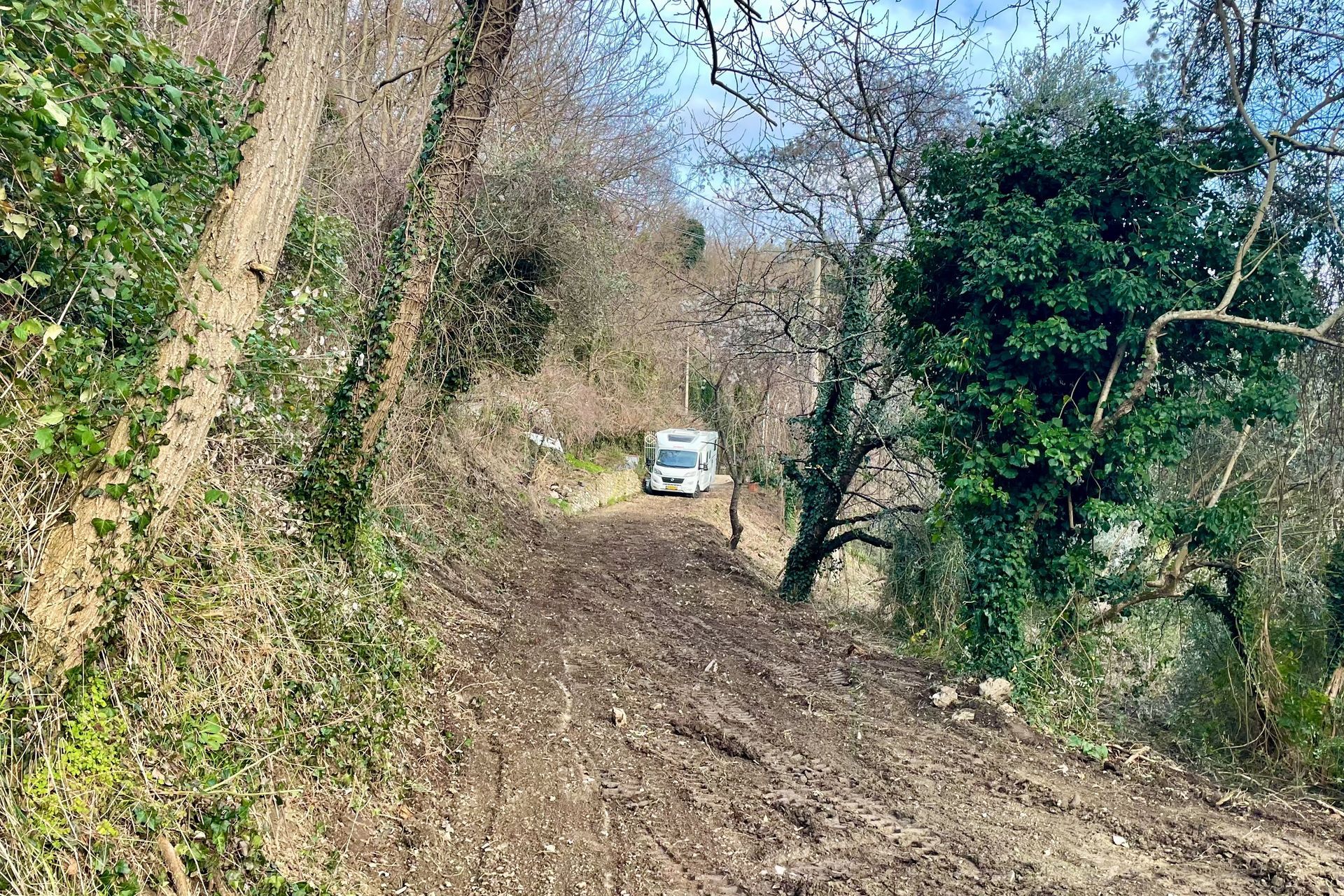 Wachten op ons plekje... Modderige weg met een witte camper, omgeven door bomen en groen.