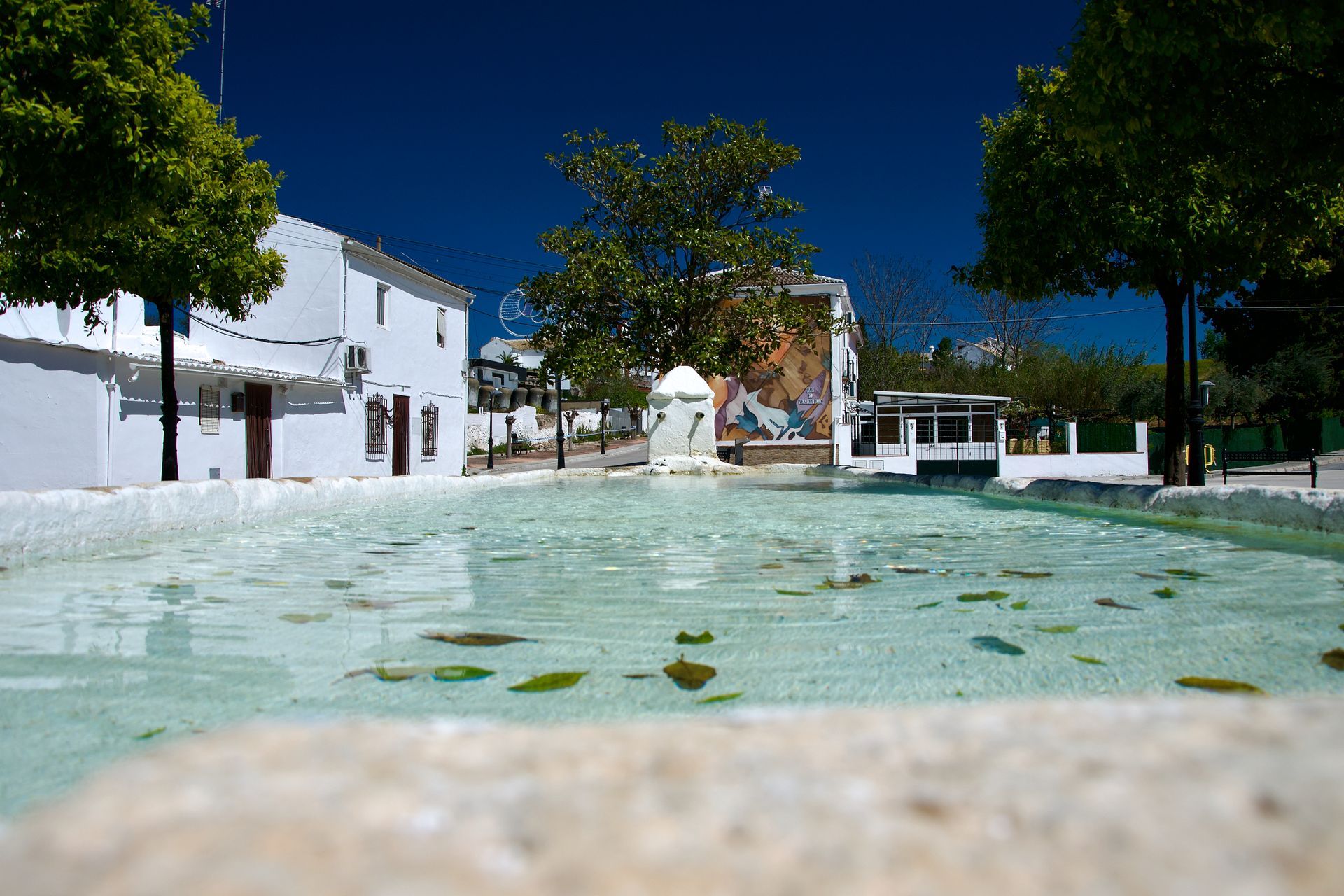 Een heldere vijver weerspiegelt de lichtblauwe lucht op een zonovergoten stadsplein, geflankeerd door witte gebouwen en groene bomen.