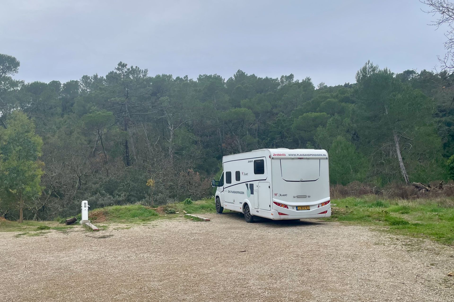 Witte camper geparkeerd op een grindpad met een bos op de achtergrond, bewolkte dag.