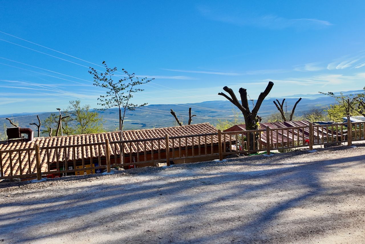 Een zonnig, verhoogd uitzicht op een rustiek gebouw met een pannendak achter een houten hek, met uitzicht op een heuvelachtig landschap.