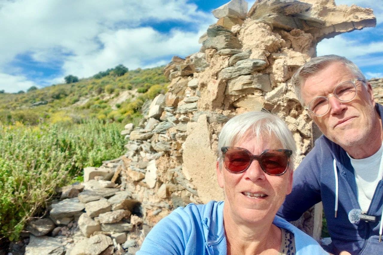 Een stel poseert voor een selfie voor een rustieke stenen ruïne op een grasrijke, zonovergoten heuvel onder een bewolkte blauwe hemel.
