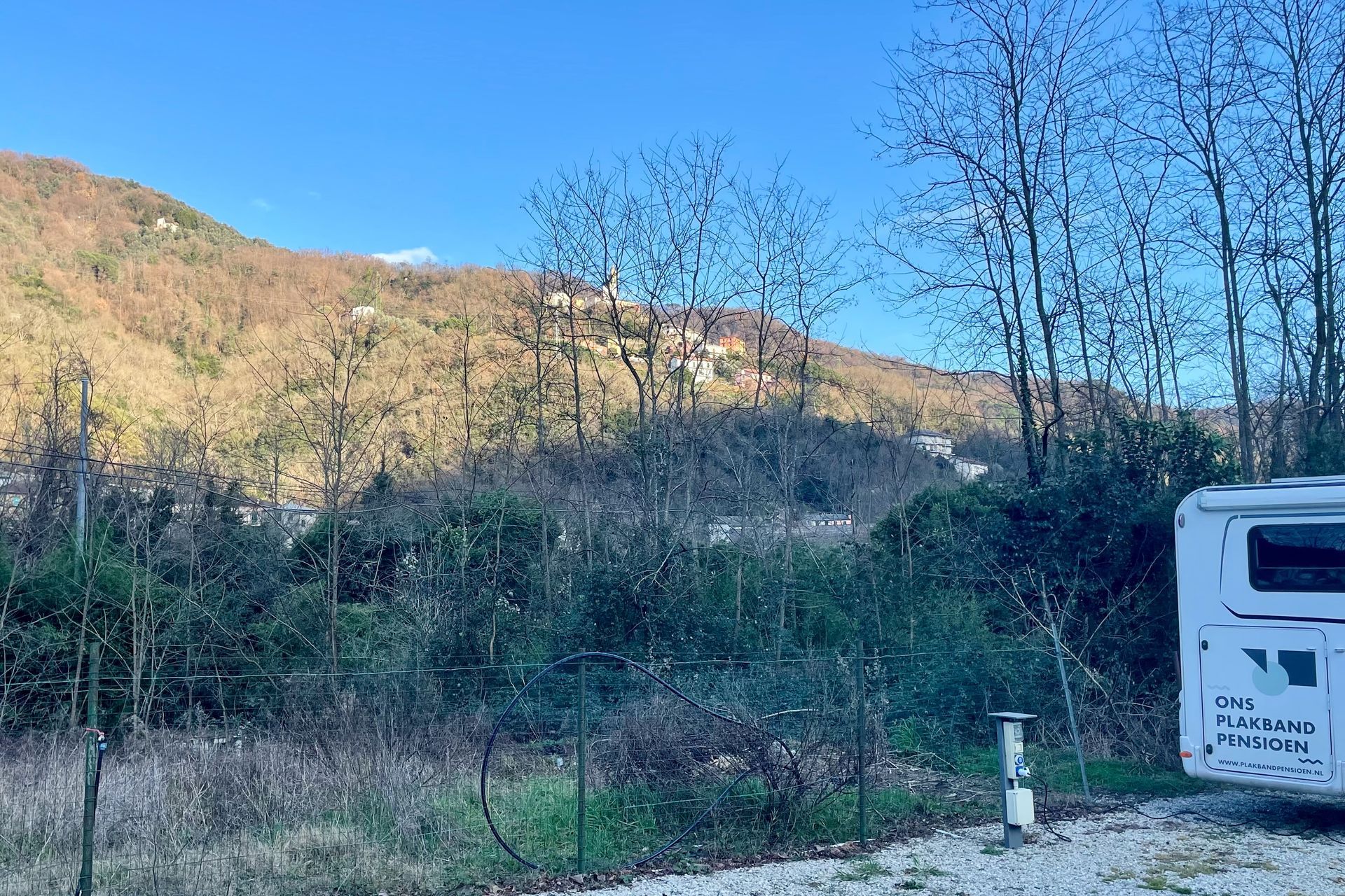 Zon op de bergen Camper geparkeerd bij een heuvel met kale bomen. Berg op de achtergrond. Groene struiken en een hek op de voorgrond.
