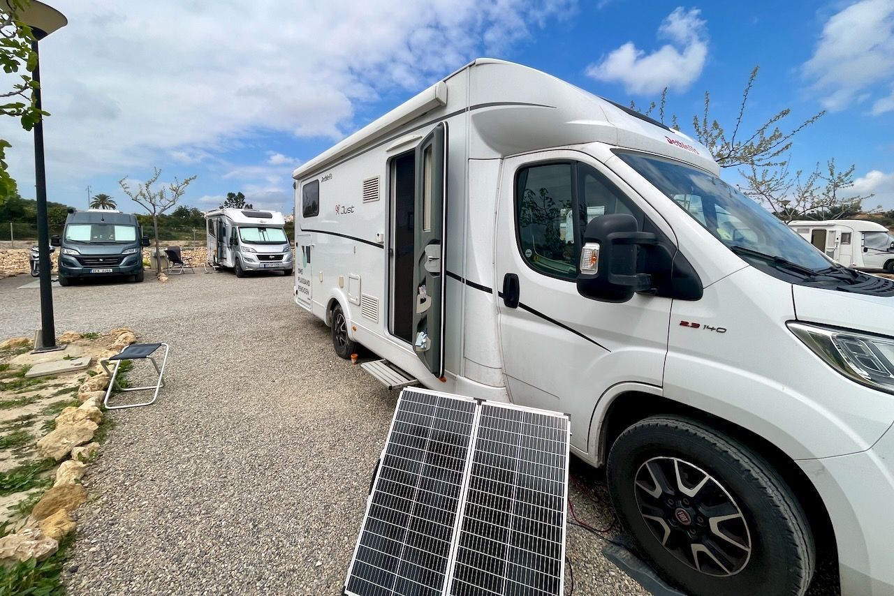 Een witte camper staat geparkeerd op een grindterrein onder een gedeeltelijk bewolkte hemel, met een draagbaar zonnepaneel ervoor opgesteld.