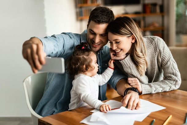 Family taking a selfie at a table while a child reaches toward papers in a bright home kitchen