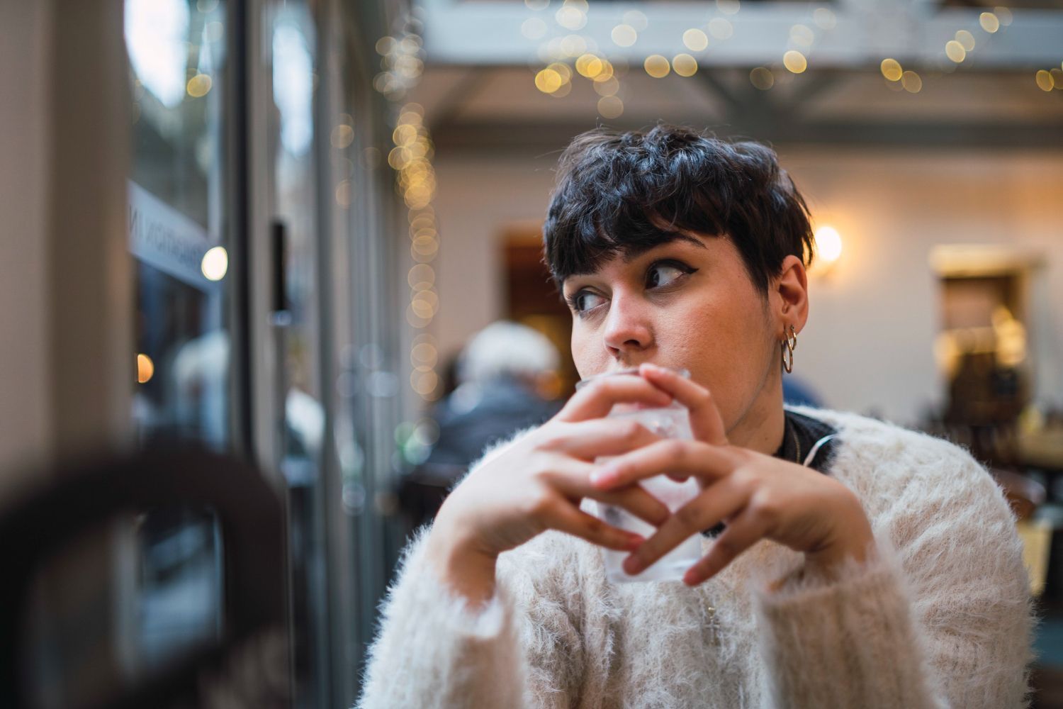 Person in a white sweater sitting in a café, hands clasped, with warm lights and blurred background.