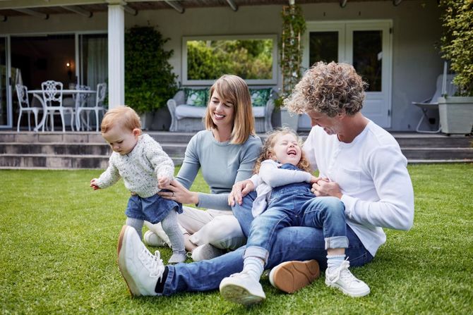 Family sitting on grass outside a house, smiling and playing with a baby and toddler