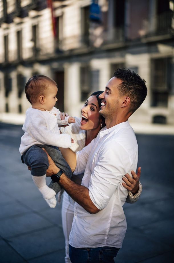 Smiling adult holding a baby outdoors on a city street, with a blurred building background