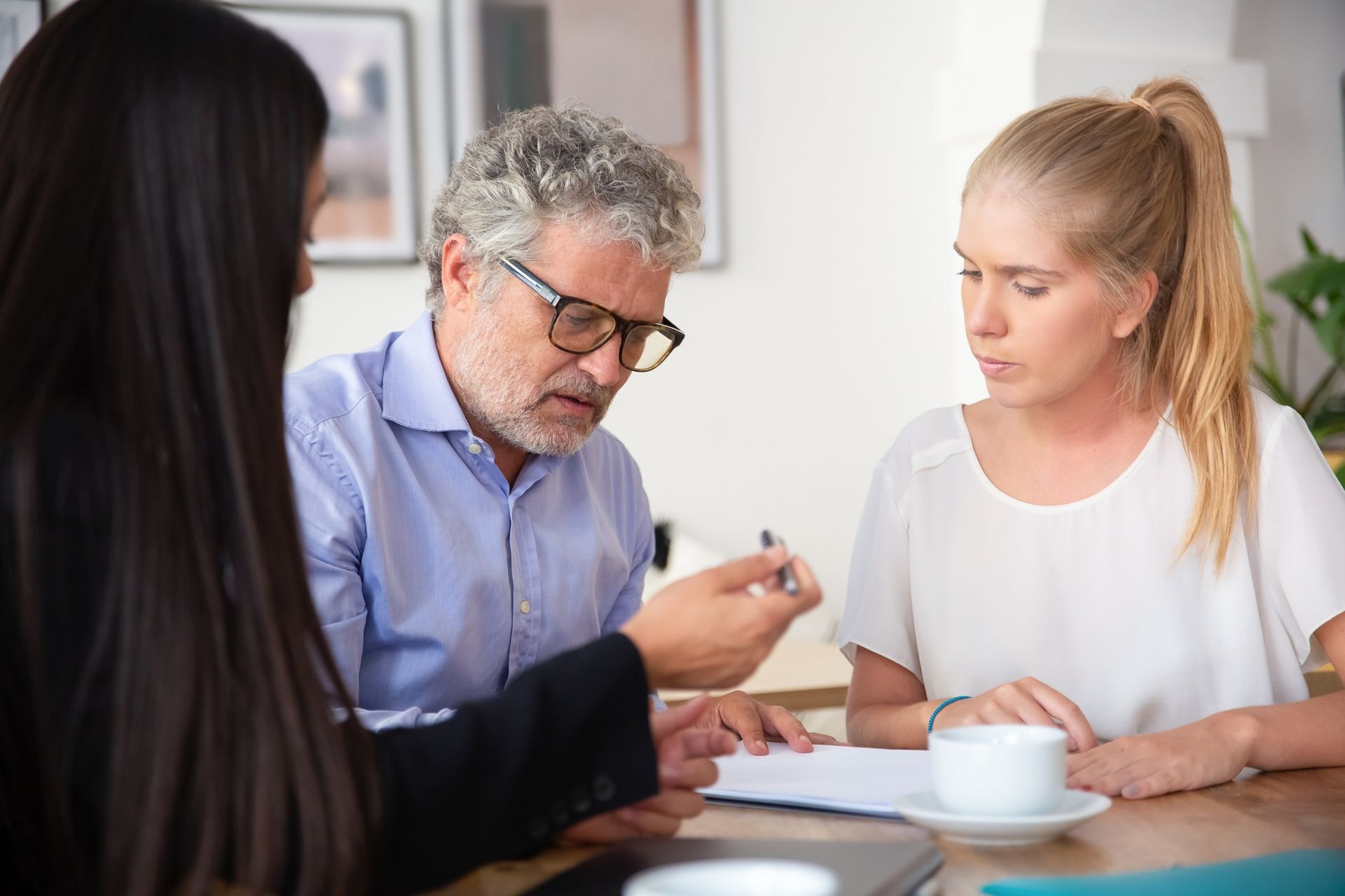 Three people discuss documents at a table in a bright office, with a cup of coffee nearby.
