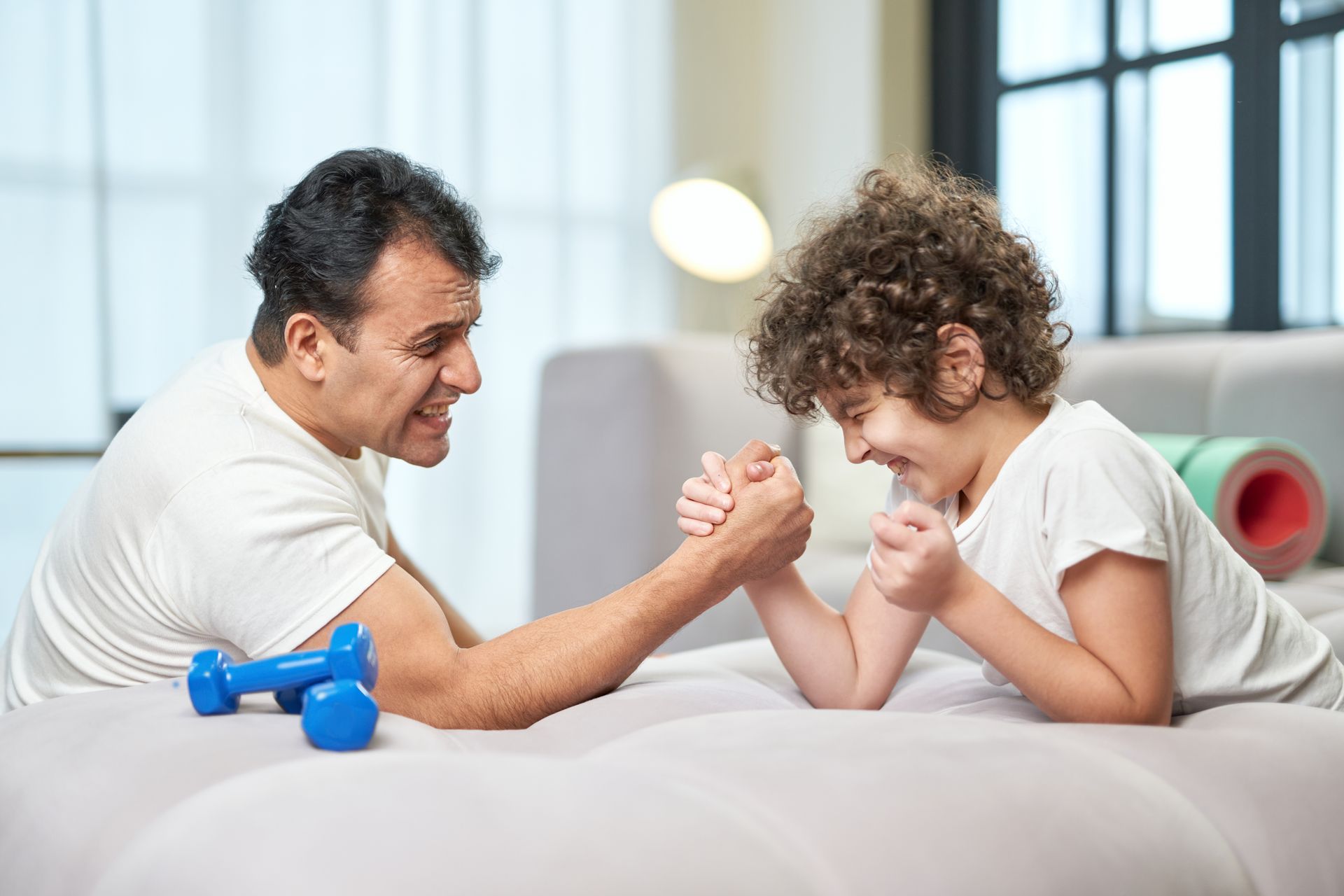 Two people arm wrestle on a couch, with blue dumbbells nearby and yoga mats in the background.
