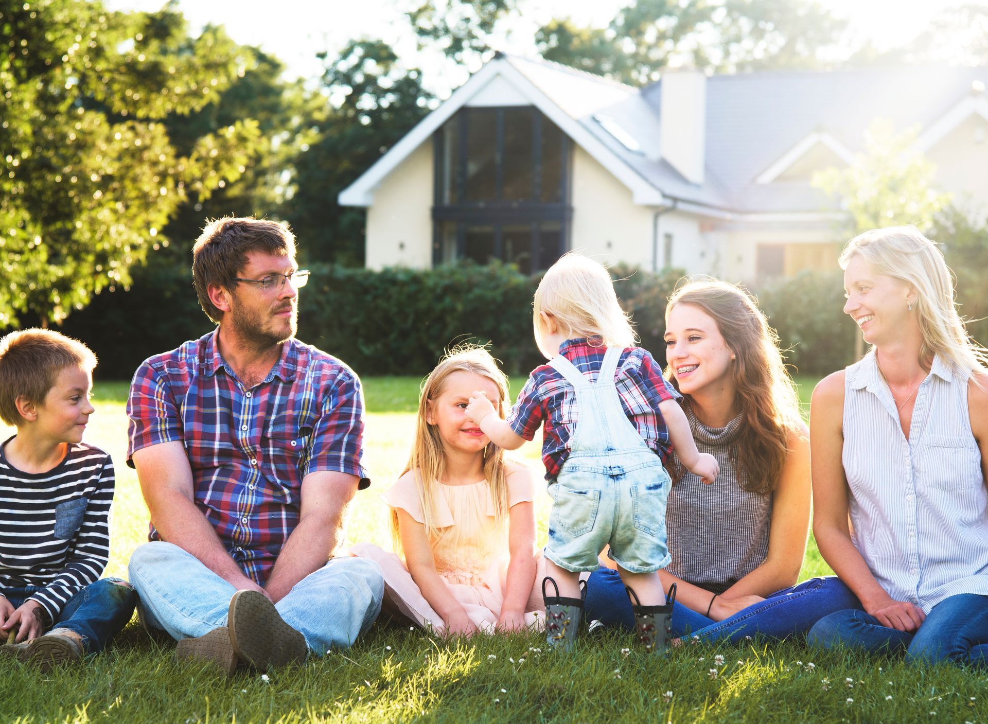 Family sitting on grass in a sunny yard, smiling and chatting in front of a house