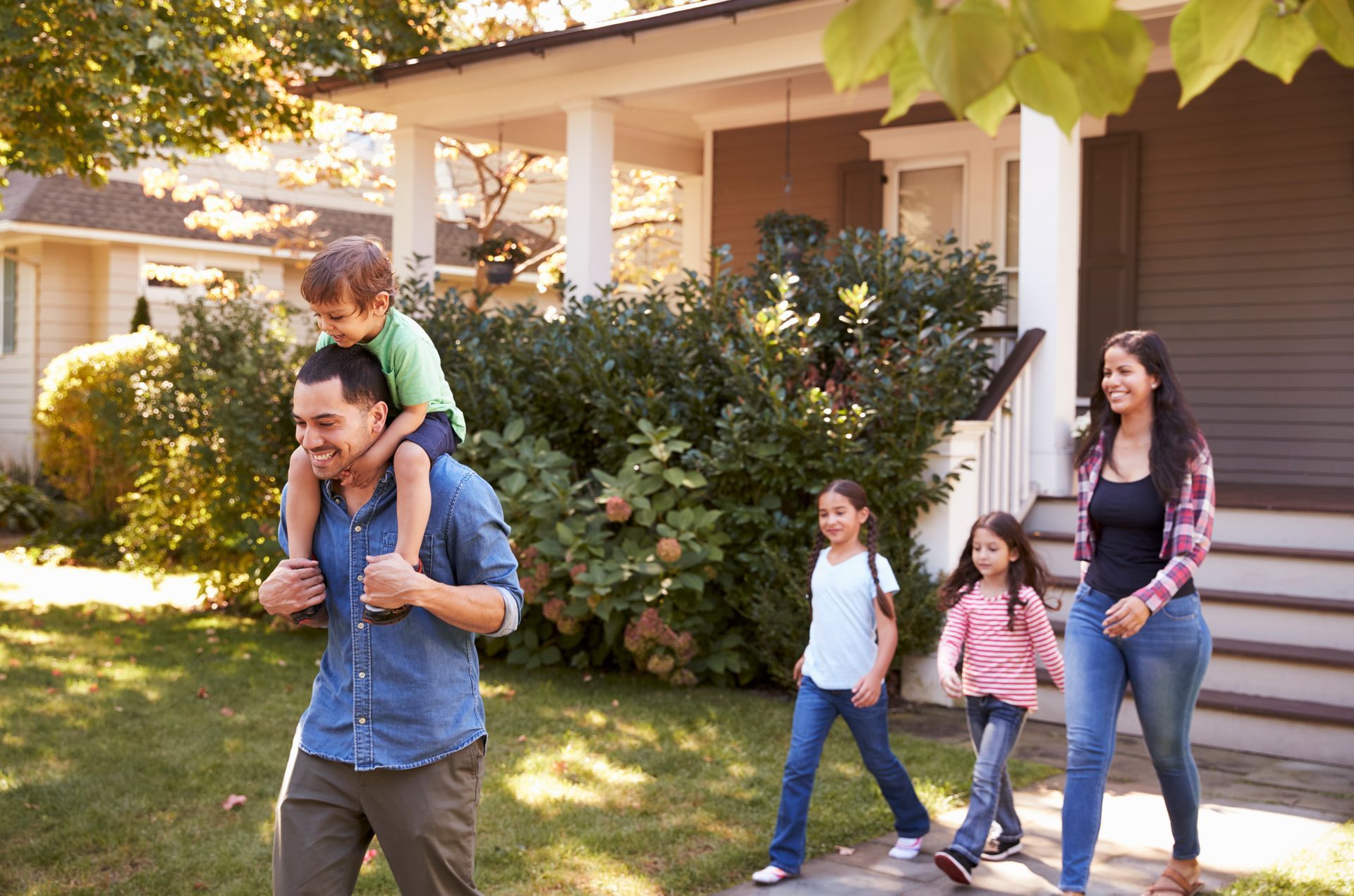 Family walking past a house, with a child riding on a man's shoulders in a sunny yard.