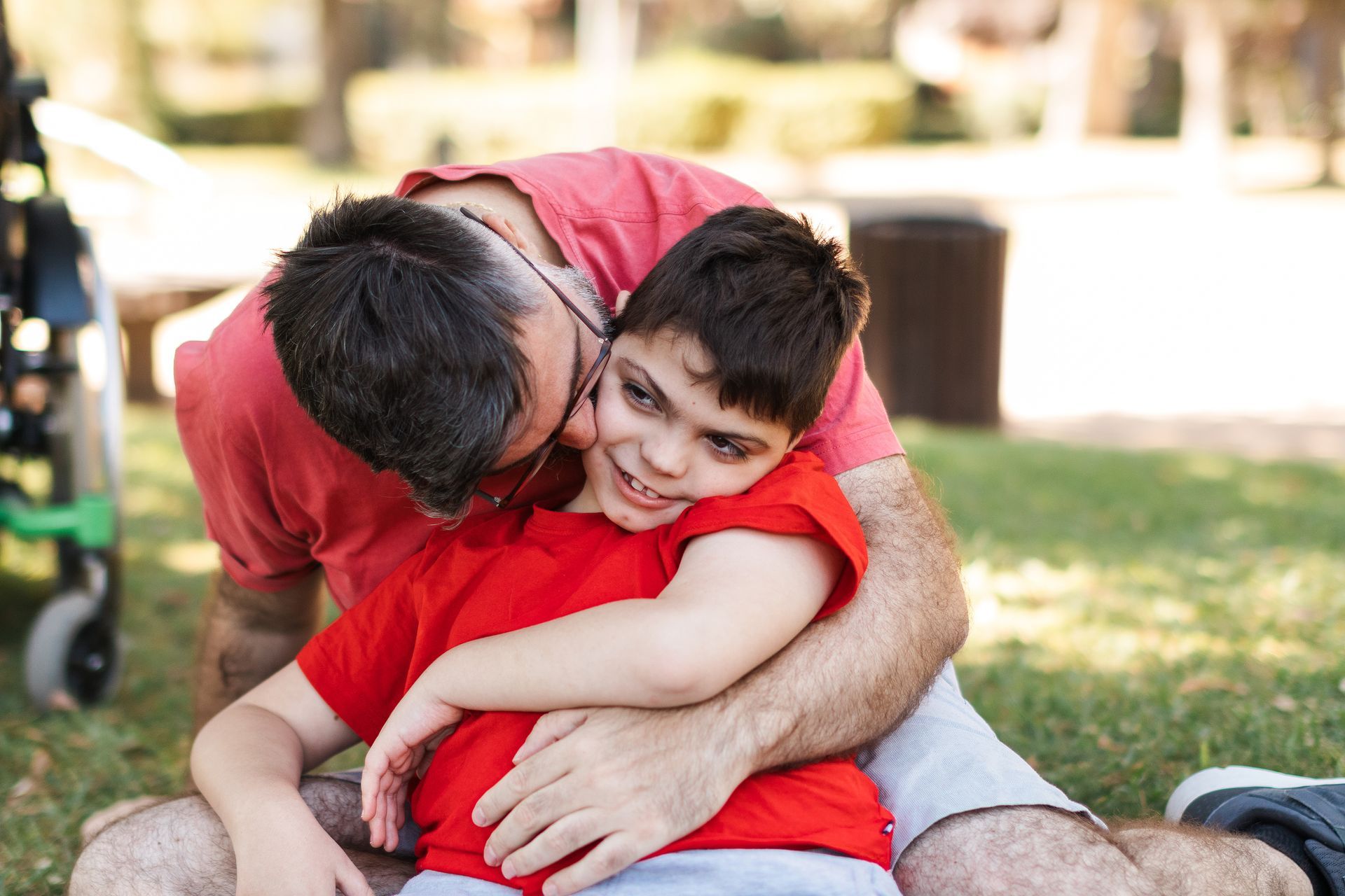 Two people hugging on grass, one in a red shirt smiling while being embraced outdoors.