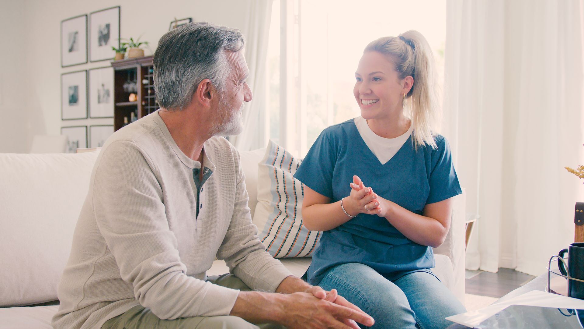 Two people smiling and talking in a bright living room, seated on a couch.